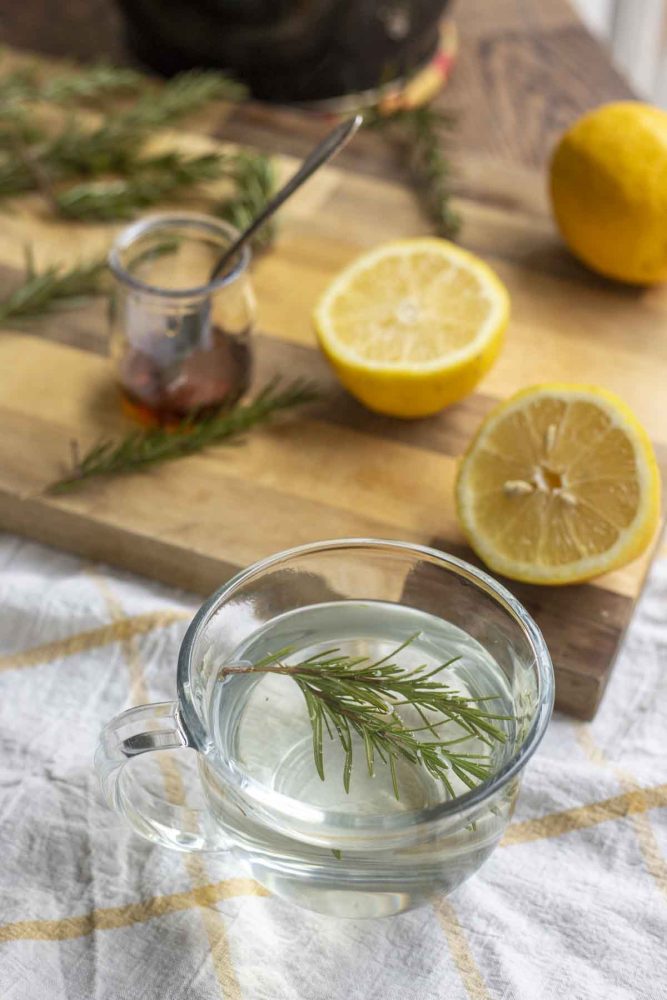glass mug of rosemary tea with a cutting board filled with sliced lemons, rosemary and a small jar of honey