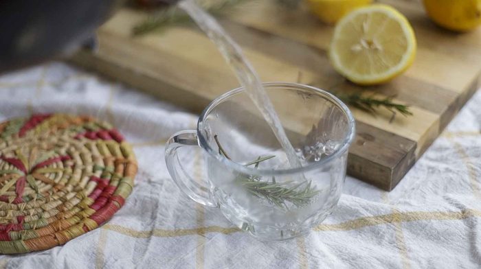 Pouring boiling water into a glass mug with rosemary sprigs. The mug sits on a yellow and white towel with a cutting board in the background
