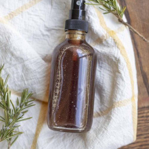 rosemary water in a spray bottle on a white and yellow towel with fresh sprigs of rosemary around the bottle