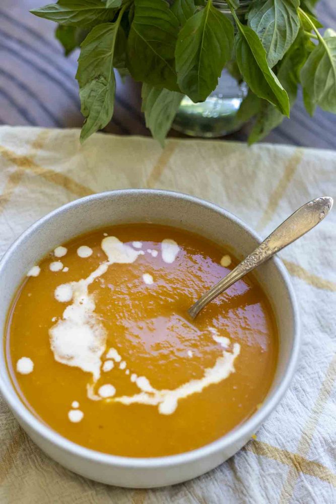 3 ingredient butternut squash soup topped with cream on top with a spoon in the soup. The bowl sits on a white and yellow towel with a bunch of basil in the background