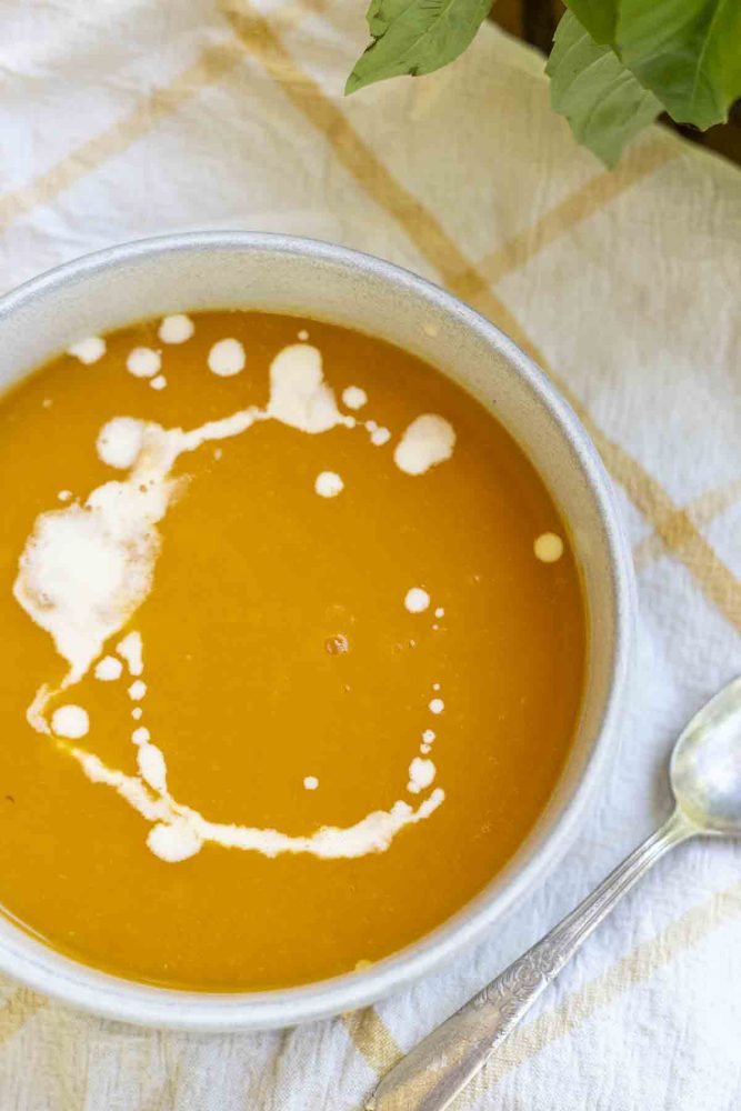 overhead photo of 3 ingredient butternut squash soup with cream drizzled on top in a bowl. The bowl is on a white and yellow check towel with a spoon to the right