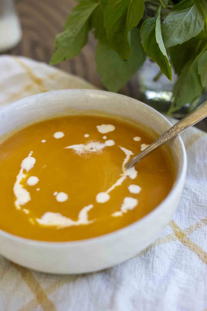 white bowl of butternut squash soup topped with a drizzle of cream and a spoon sits in the bowl. The bowl is on a white and yellow checked towel on a wood countertop with a bunch of basil in the background