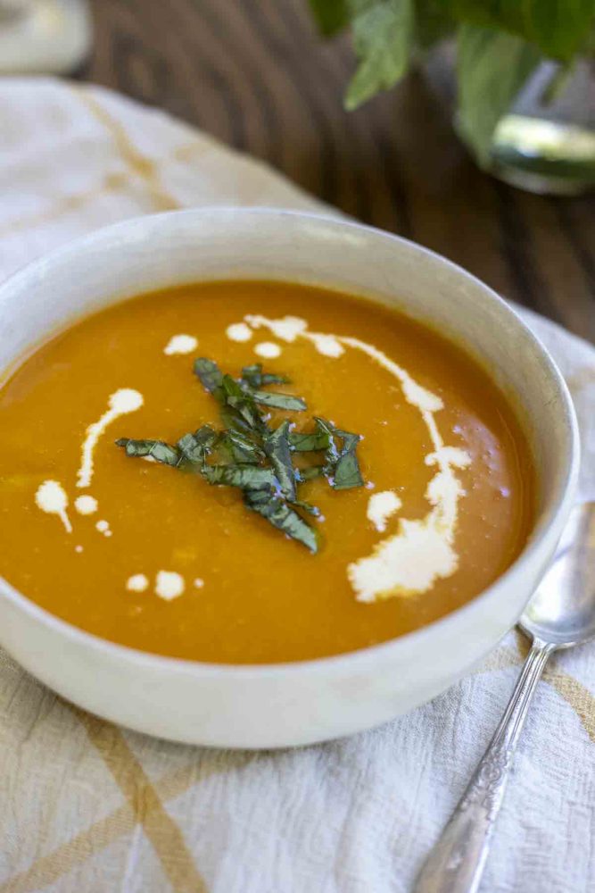 bowl of 3 ingredient butternut squash soup drizzled with cream and topped with basil in a white bowl on a white and yellow checked towel. 