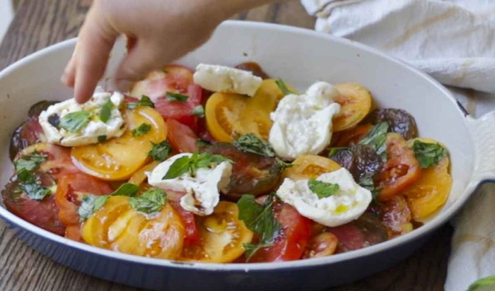 hand sprinkling basil on top of tomatoes and burrata cheese in a white and blue baking dish