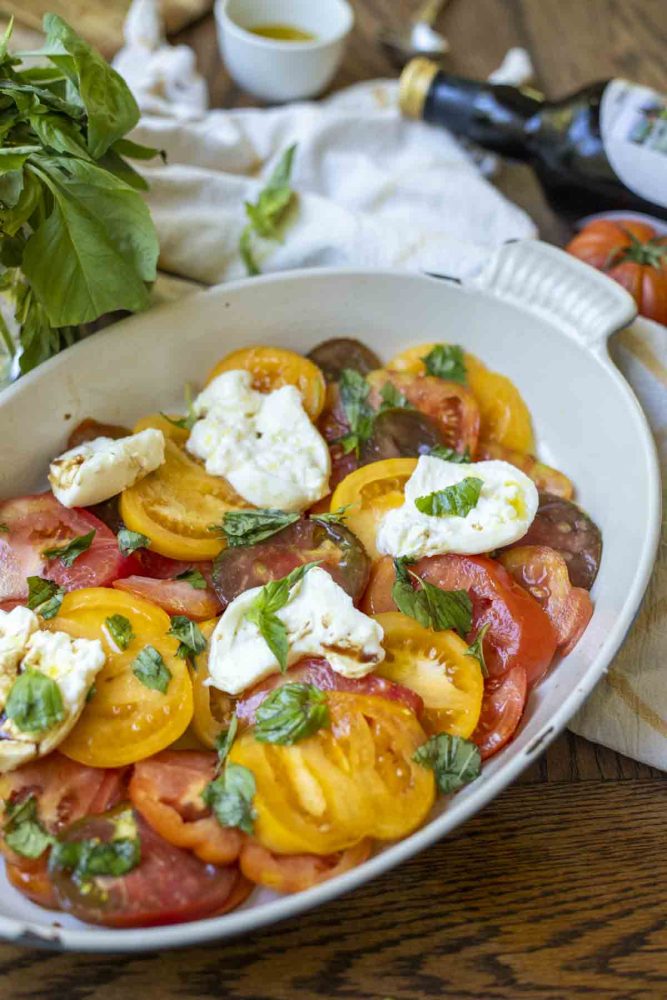 a white ceramic dish with a burrata caprese salad topped with basil. Ingredients surround the baking dish