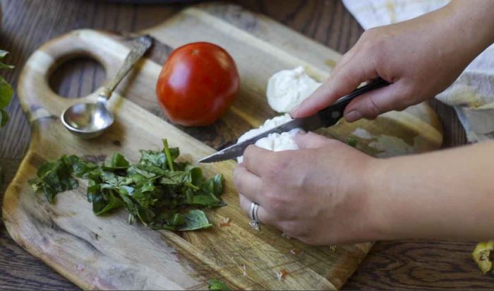 slicing burrata cheese on a cutting board with a tomato and basil surrounding the cheese
