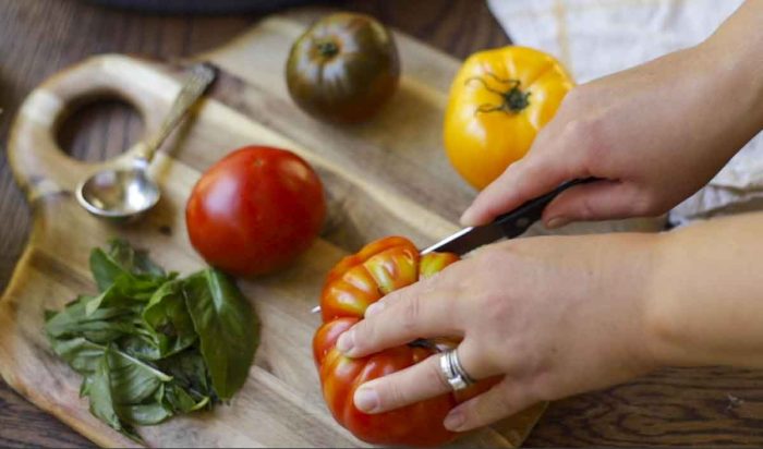 slicing tomatoes with a knife on a cutting board with a variety of colored tomatoes and fresh basil
