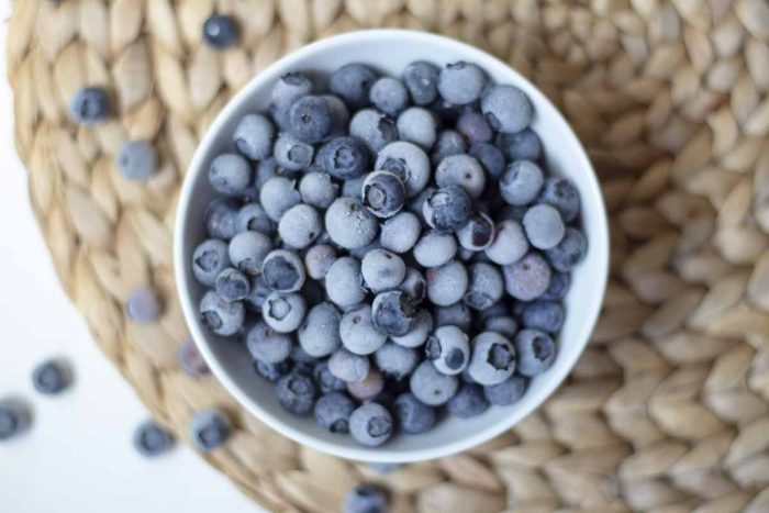 frozen blueberries in a white bowl on a woven place mat surrounded by blueberries