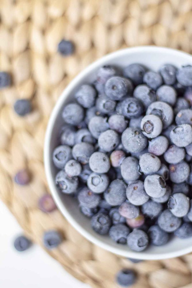 Overhead photo of frozen blueberries in a white bowl on a woven mat with more blueberries on the mat.