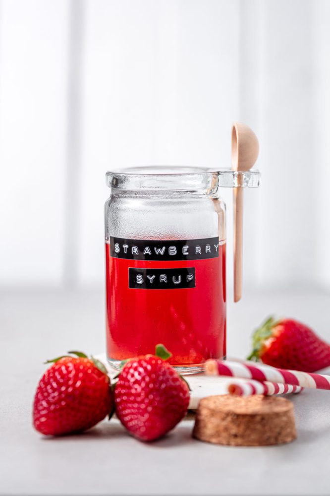 Strawberry simple syrup in a glass jar on a marble countertop with strawberries surrounding the jar