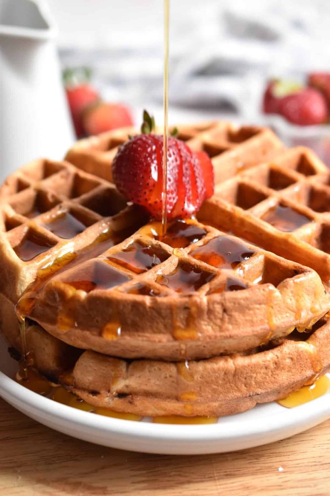 strawberry waffles topped with a strawberry as maple syrup is drizzled over top