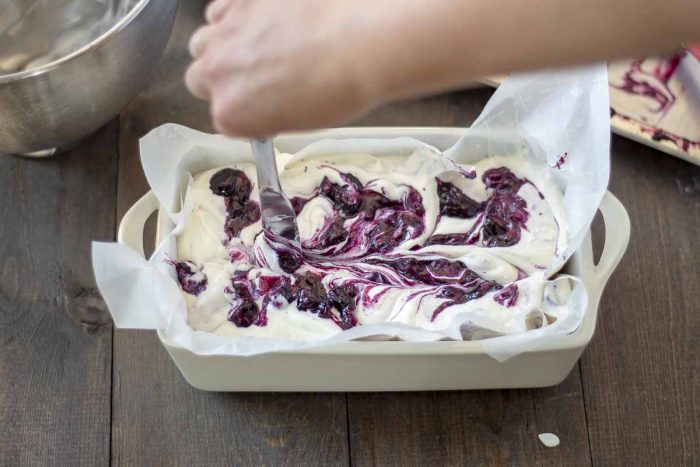 hand holding knife and creating swirls in blueberry cheesecake ice cream in a parchment lined loaf pan on a wood table