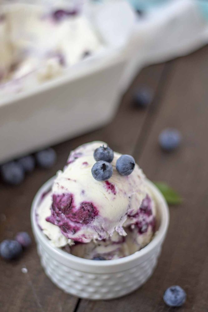 two scoops of blueberry cheesecake ice cream topped with fresh blueberries in a white dish on a wood table with blueberries around the ice cream