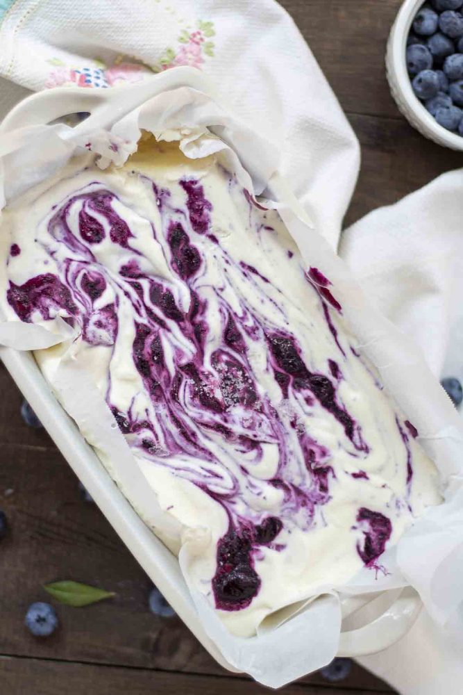 overhead photo of a loaf pan lined with parchment paper full of blueberry cheesecake ice cream with swirls of blueberries. The pan is on a white towel with blueberries all around