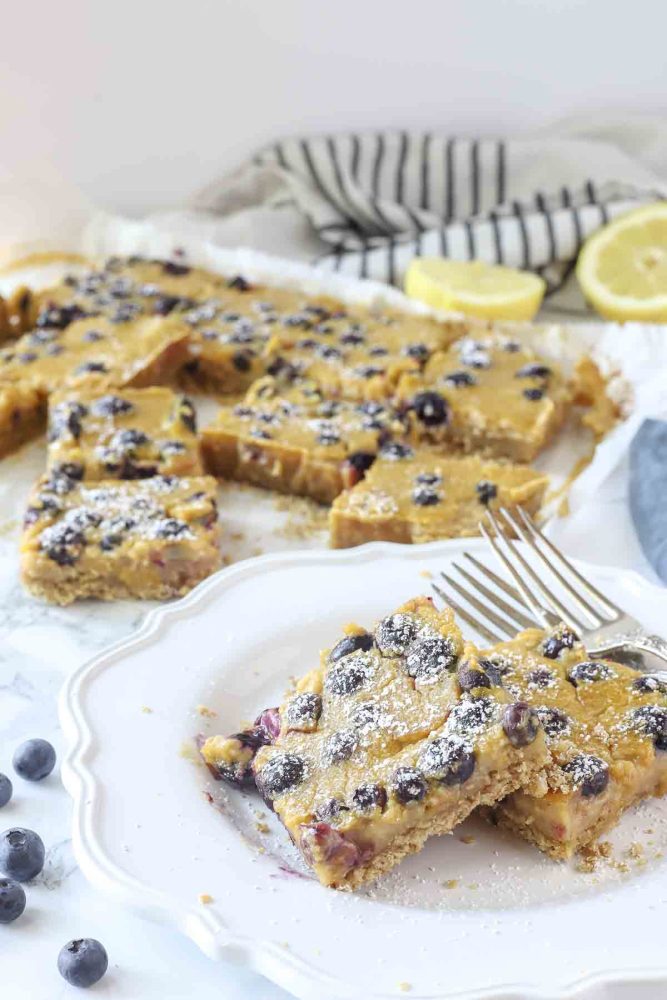two blueberry lemon pie bars on a white plate with more bars on parchment paper in the background