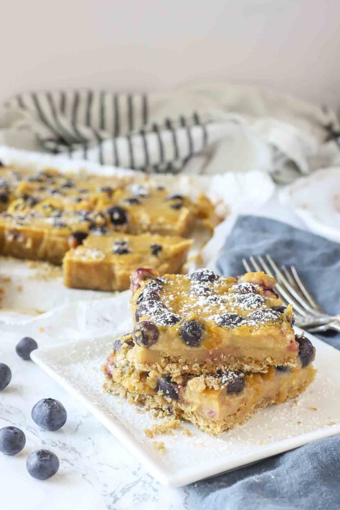 Two blueberry lemon bars stacked on a white place with a blue napkin to the right