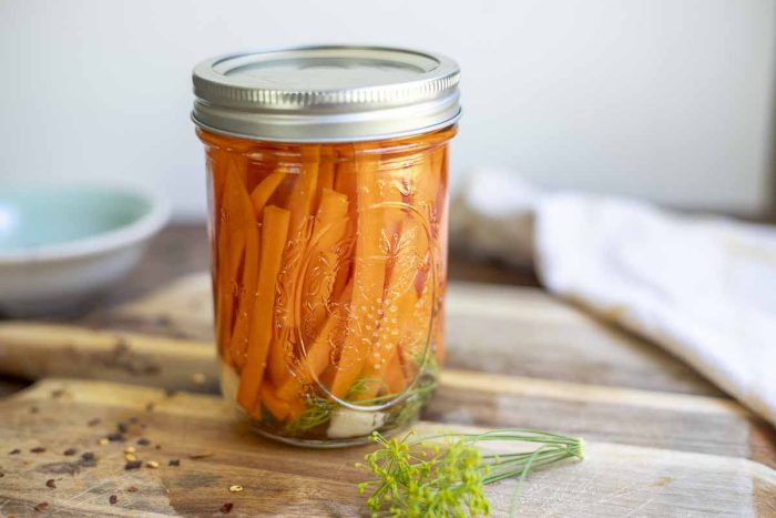 jar of pickled carrots on a wood cutting board with dill and red pepper flakes. A teal bowl is in the background