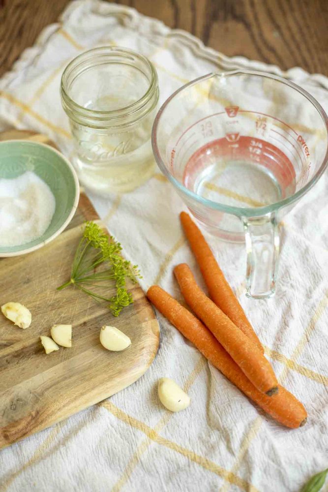 carrots, vinegar and water in jars, a dish of salt, garlic, and fresh dill on a cutting board
