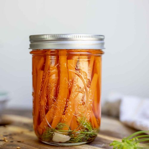 jar of quick pickled carrot sticks with garlic and dill on a wood cutting board with fresh dill