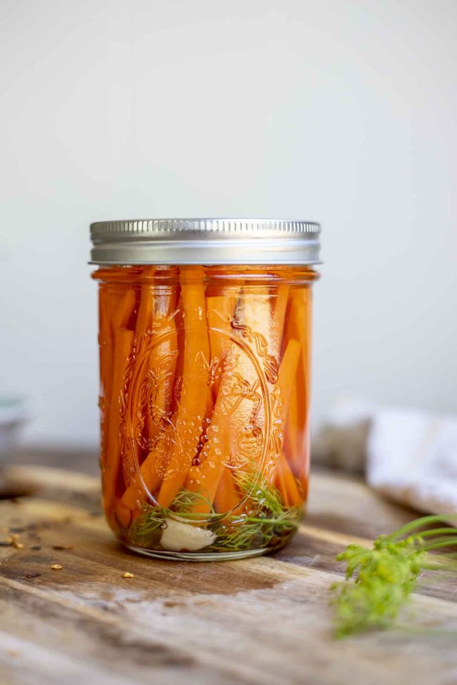 jar of quick pickled carrot sticks with garlic and dill on a wood cutting board with fresh dill