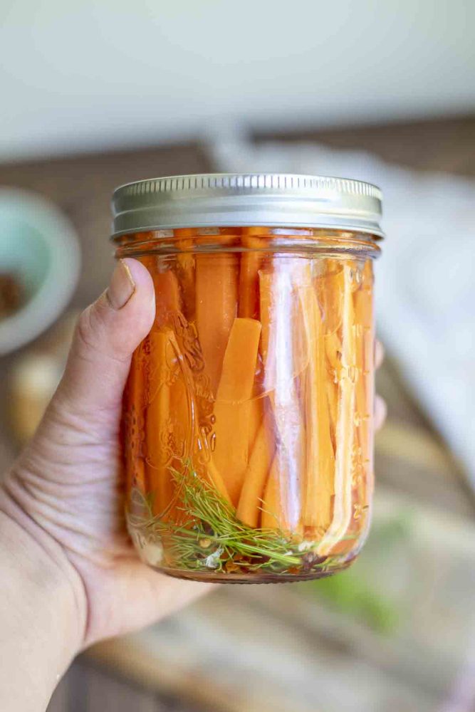 hand holding a jar of quick pickled carrots with dill and garlic. A wood cutting board is in the background