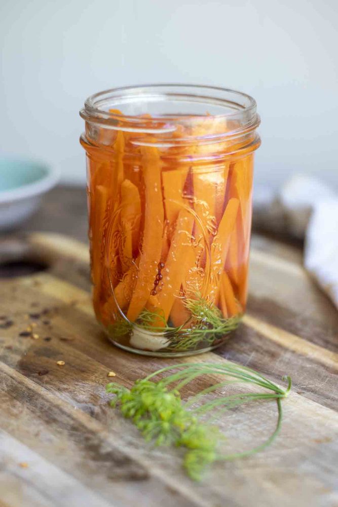 open jar of carrots in a brine with dill and garlic on a wood cutting board with fresh dill and red pepper flakes