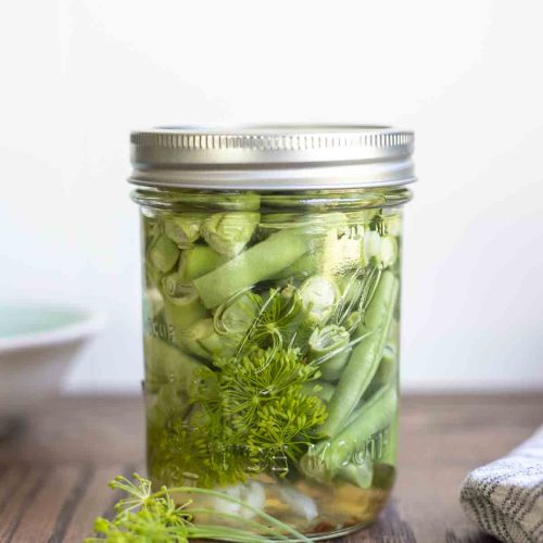 pickled green beans in a mason jar with a lid on a wood table. Fresh dill sits in front of the jar