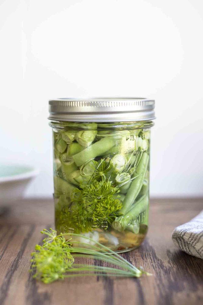 pickled green beans in a mason jar with a lid on a wood table. Fresh dill sits in front of the jar