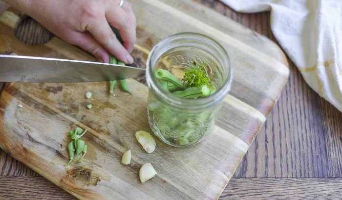 cutting green beans with a knife on a wood cutting board. A jar with garlic and dill also sit on the cutting board