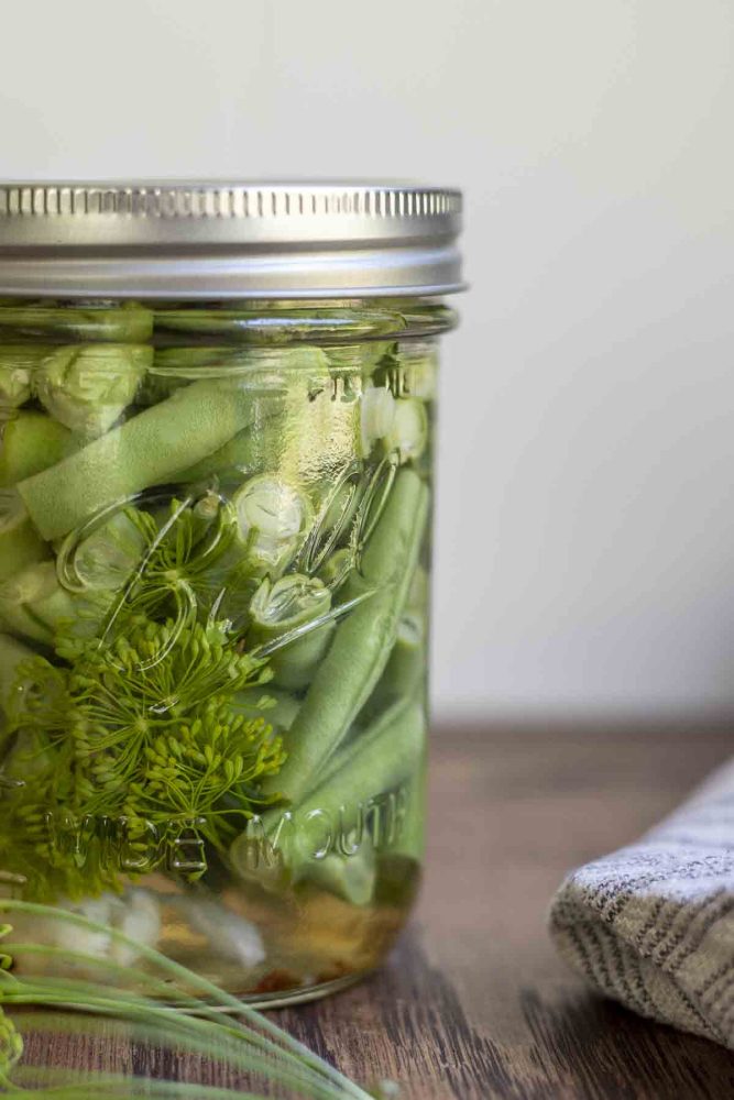 up close picture of quick pickled green beans on a wood table. a fresh head of dill in front of the jar