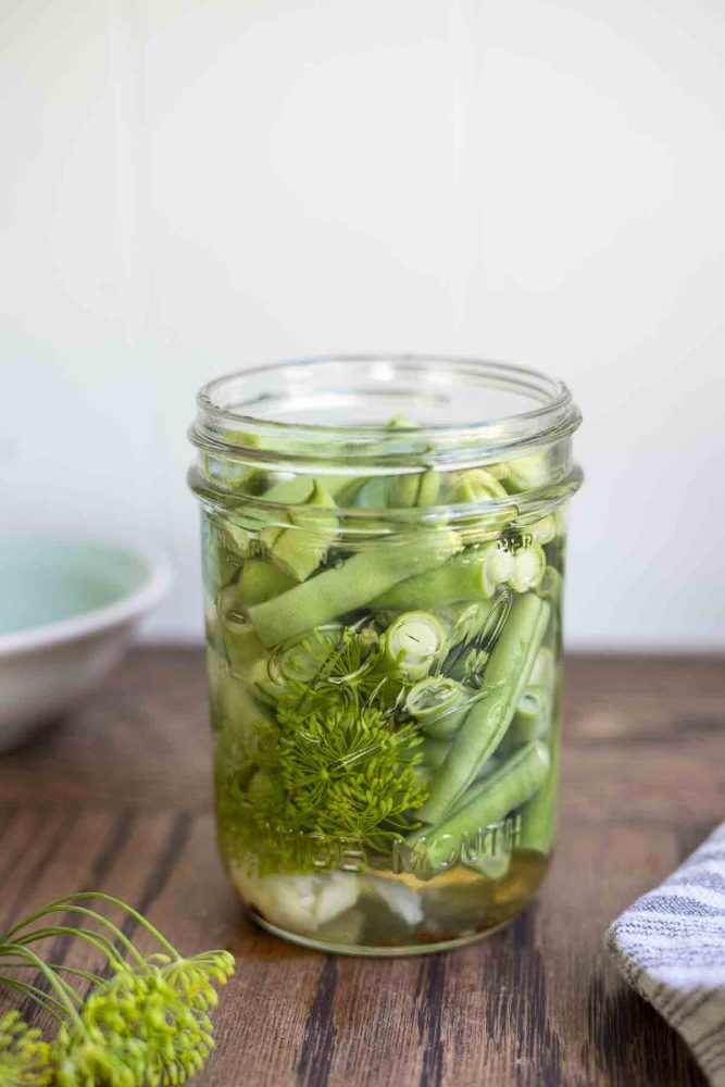 a mason jar of pickled green beans with garlic and dill on a wood table with fresh dill in front and a teal bowl in the background