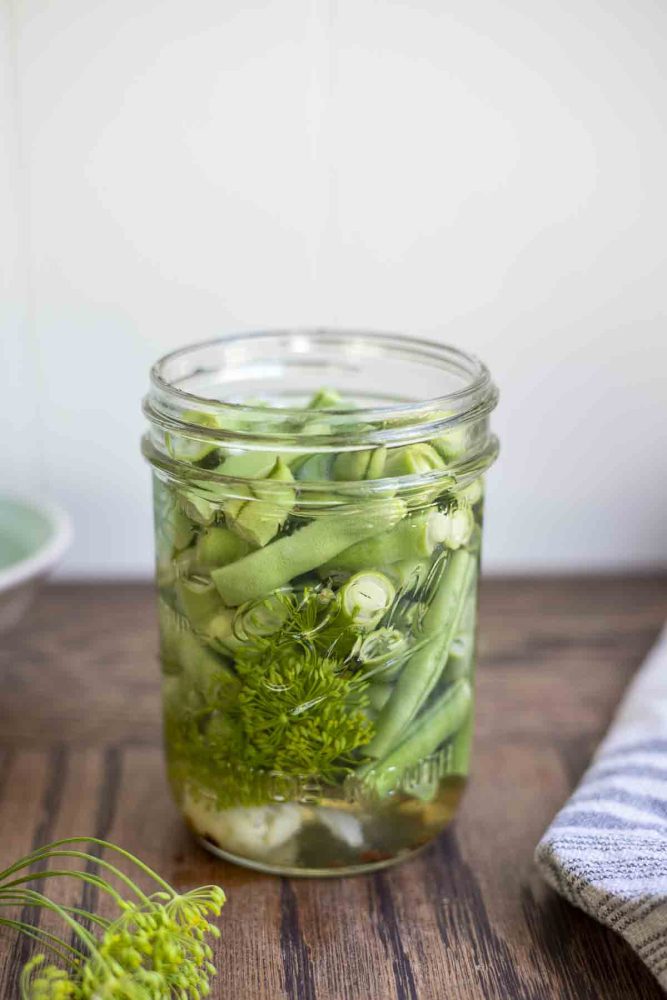 a open jar of pickled green beans with fresh dill, garlic, and red pepper flakes on a wood table. Fresh dill is in front of the jar