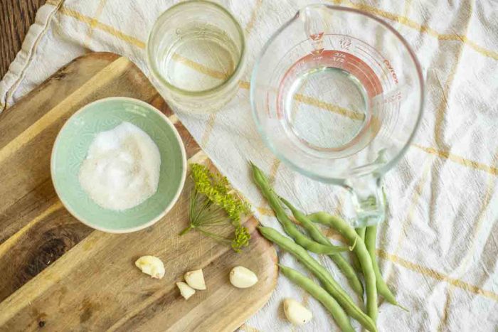 containers or vinegar, water, salt and sugar, and green beans, garlic and dill on a cutting board