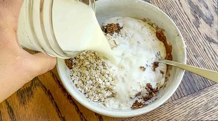 milk being poured into a bowl with mashed banana, oats, pumpkin puree, chia seeds, and pumpkin spice