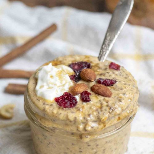 healthy pumpkin pie overnight oats topped with yogurt, brown sugar, almonds, and dried cranberries on a white and yellow towel with cinnamon sticks in the background