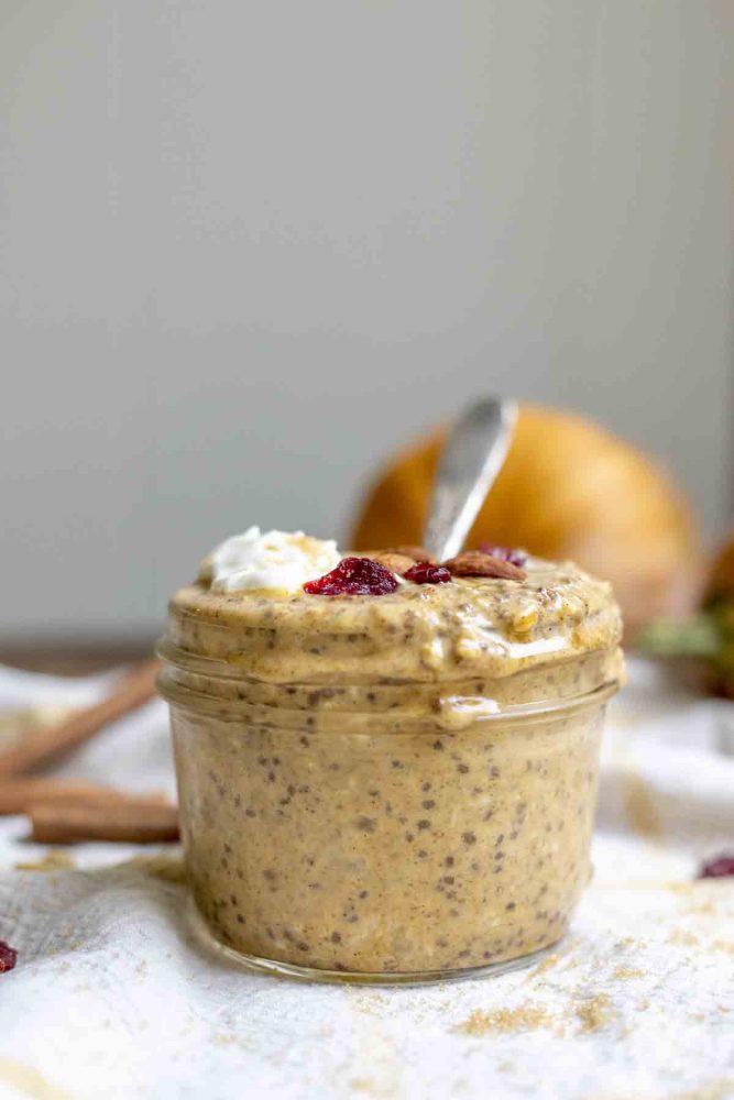 side view of pumpkin spice overnight oats in a small mason jar. The oats are topped with yogurt, brown sugar, cranberries, and almonds. The jar sits on a white and yellow towel with cinnamon sticks and pumpkins in the background