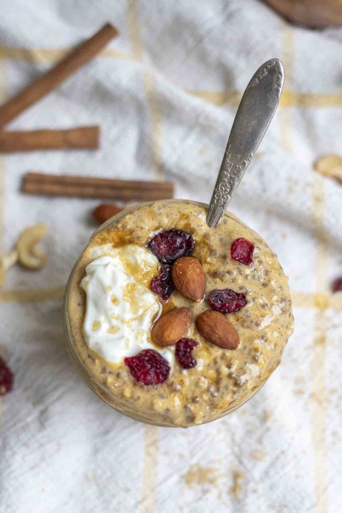 overhead photo of a small mason jar with pumpkin pie overnight oats topped with yogurt, cranberries, and almonds with a spoon in the jar. There is a spoon in the overnight oats. The jar sits on a white and yellow towel with cinnamon sticks and nuts surrounding the jar