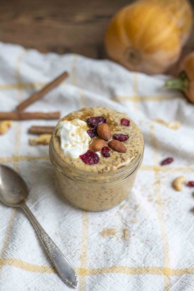 jar of pumpkin pie overnight oats topped with brown sugar, yogurt, cranberries, and almonds on a white and yellow towel. A spoon sits to the left and the jar is surrounded by pumpkins, cinnamon sticks, nuts and cranberries