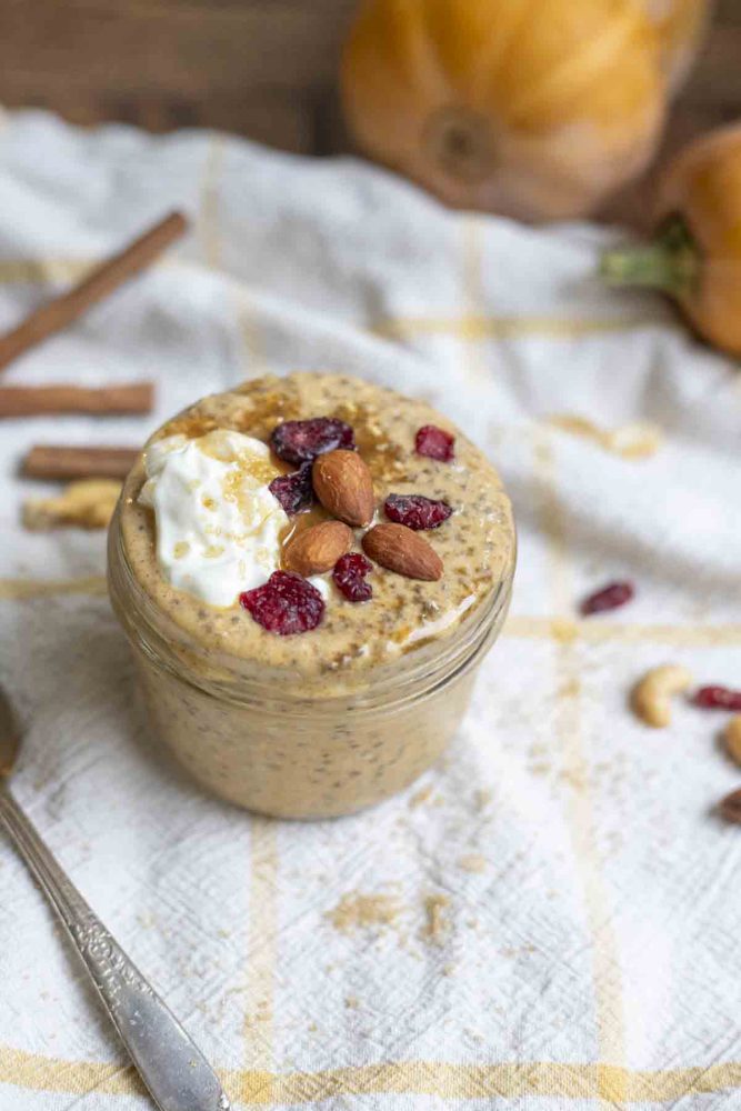 jar of pumpkin spice overnight oats toped with a dollop of yogurt, cranberries, and almonds on a white and yellow towel. A spoon, cinnamon sticks, dried cranberries and nuts are surrounding the jar