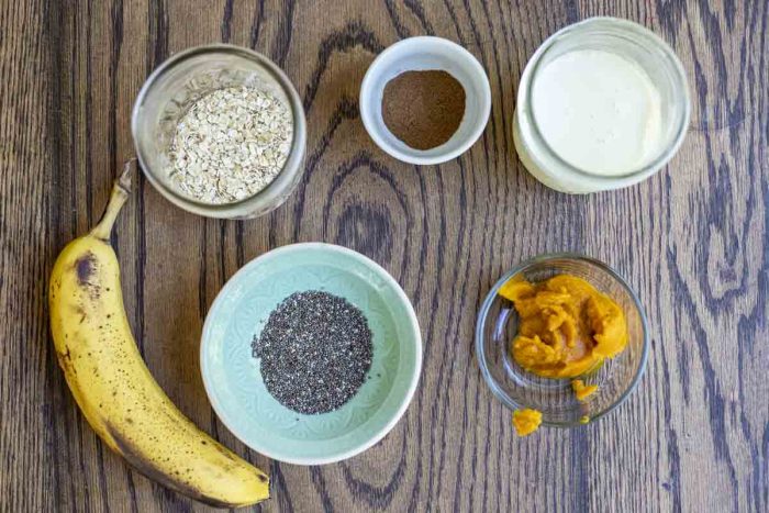 jars of oats, milk, pumpkin spice, canned pumpkin, chia seeds and a banana on a wood countertop