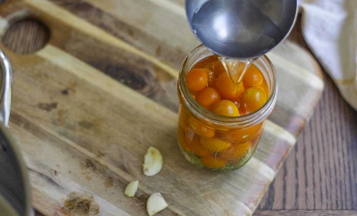 pouring a brine into a mason jar of cherry tomatoes on a wood cutting board with garlic in front of the jar