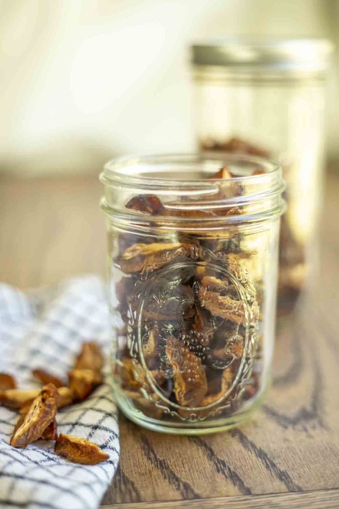 jar of dried plums on a wood table with another jar in the background. A black and white towel will more prunes are to the right