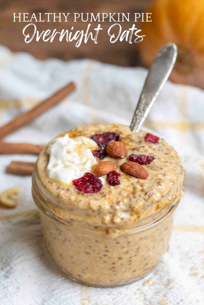 healthy pumpkin pie overnight oats topped with yogurt, brown sugar, almonds, and dried cranberries on a white and yellow towel with cinnamon sticks in the background