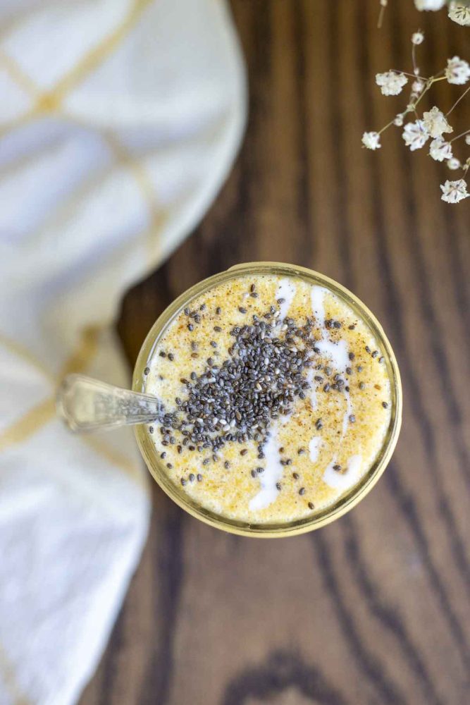 overhead photo of a pumpkin smoothie topped with yogurt and chia seeds in a glass jar on a wood countertop. There is a white and yellow towel to the left