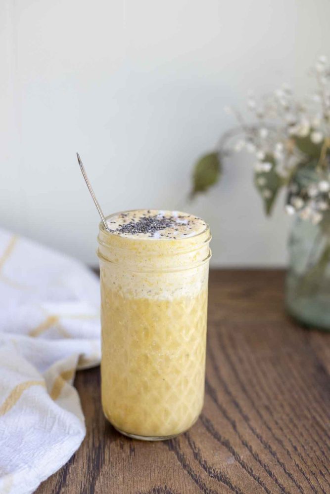 healthy pumpkin smoothie in a glass jar with a spoon in the smoothie. The jar is on a wood countertop with a white and yellow towel and a jar of flowers int he background