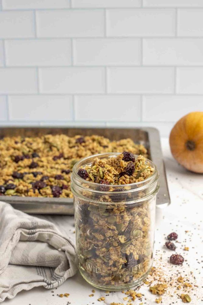 a jar of pumpkin granola with dried cranberries in a mason jar on a marble countertop with oats, chia seeds, and cranberries surrounding the jar. A pan of more granola and a pumpkin is in the background