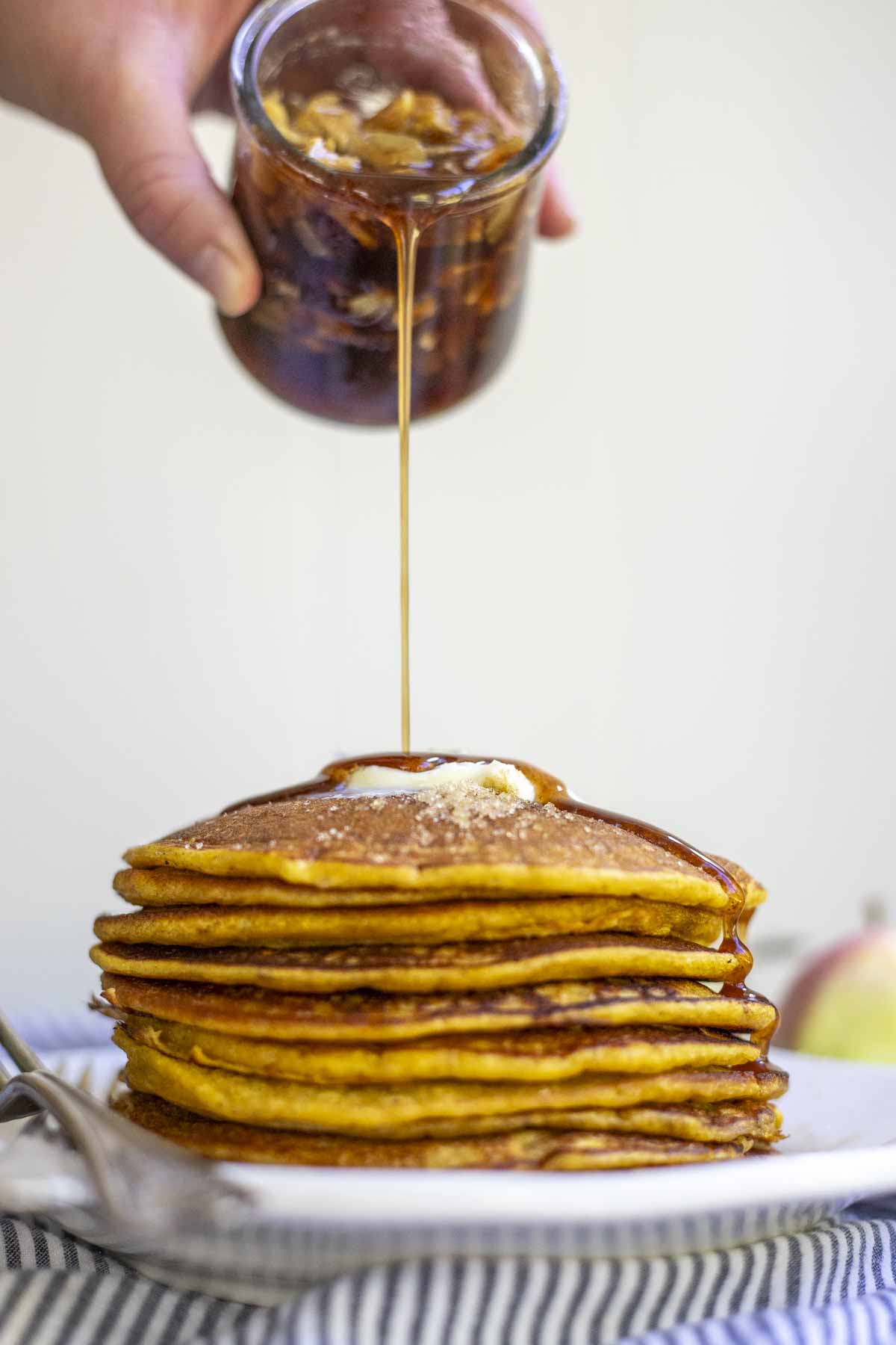 apple syrup being poured over a stack of pumpkin pancakes with butter on a white plate.