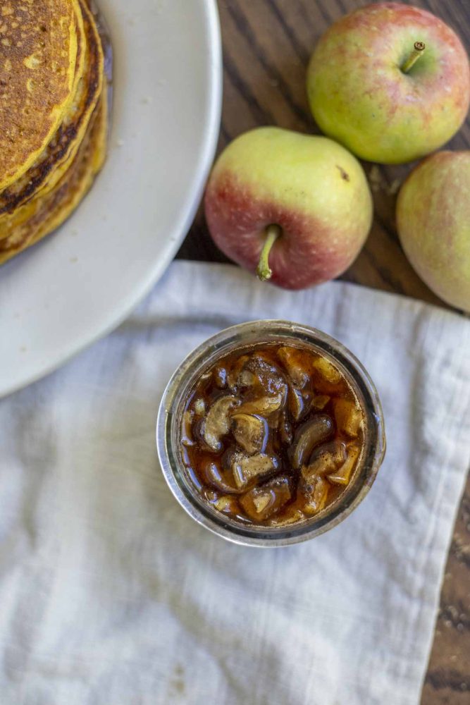 overhead photo of apple syrup in a small jar on a tan napkin with apples and pancakes in the background