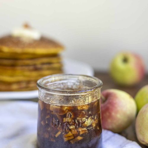cinnamon apple syrup in a small glass jar on a napkin with apples and a stack of pumpkin pancakes in the background