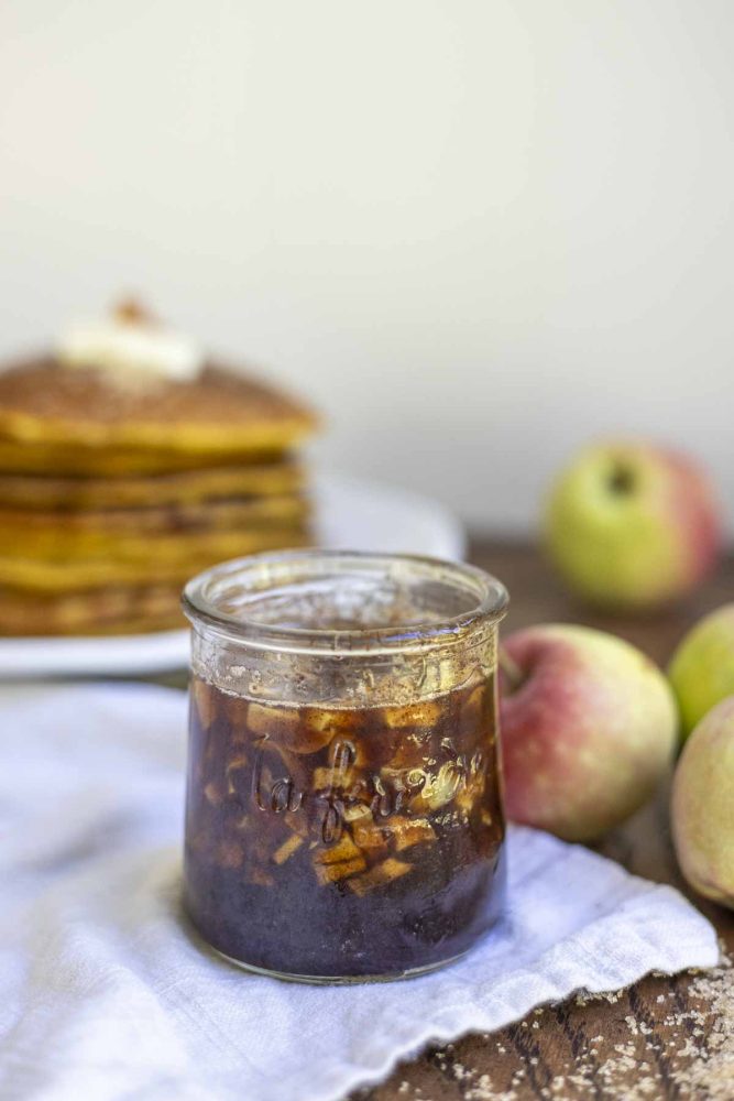cinnamon apple syrup in a small glass jar on a napkin with apples and a stack of pumpkin pancakes in the background